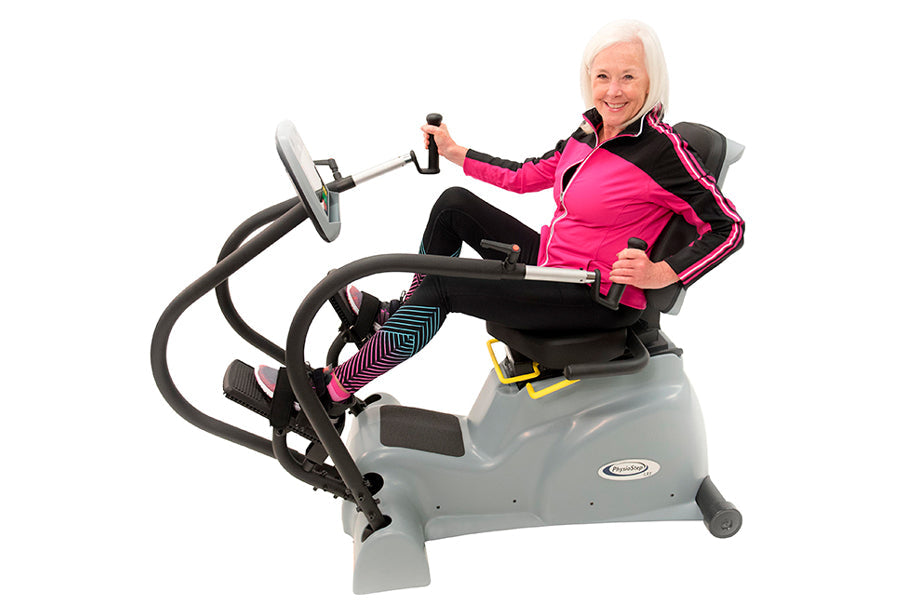 Woman wearing hot pink and black athletic jacket demonstrating gray Physiostep LXT recumbent elliptical cross trainer with bl