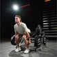 Man wearing light gray t-shirt performing deadlift on Hammer Strength squat machine with loaded barbell in gym setting with n