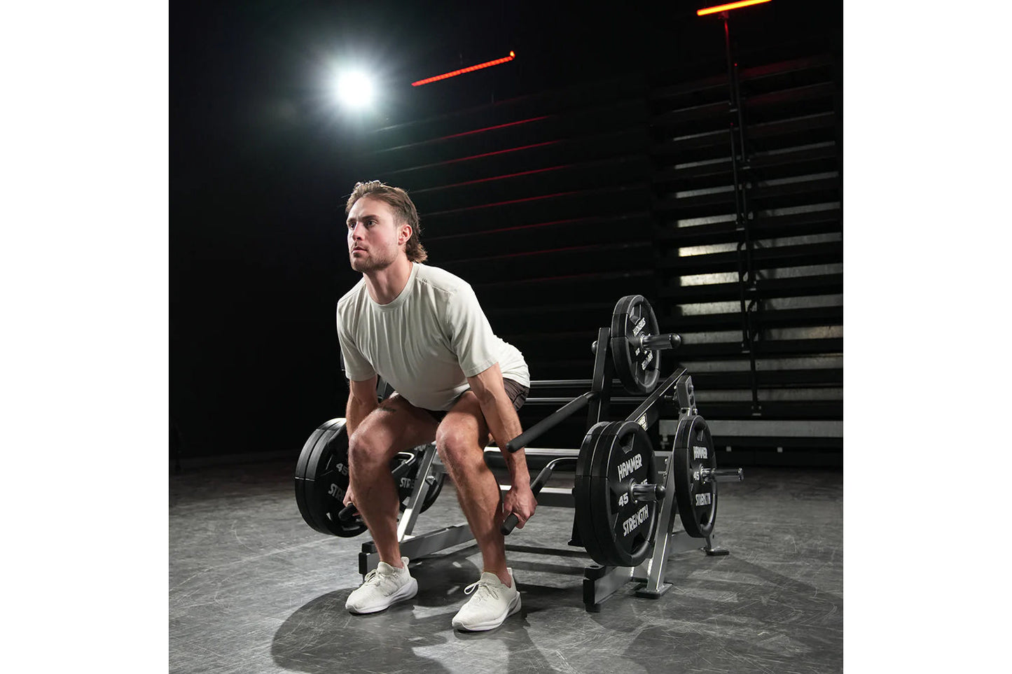 Man wearing light gray t-shirt performing deadlift on Hammer Strength squat machine with loaded barbell in gym setting with n