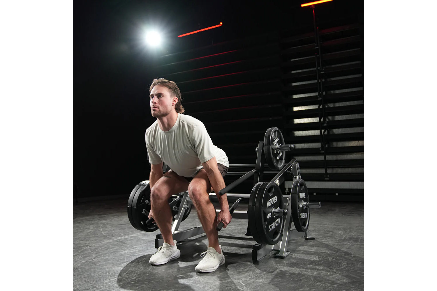 Man wearing light gray t-shirt performing deadlift on Hammer Strength squat machine with loaded barbell in gym setting with n