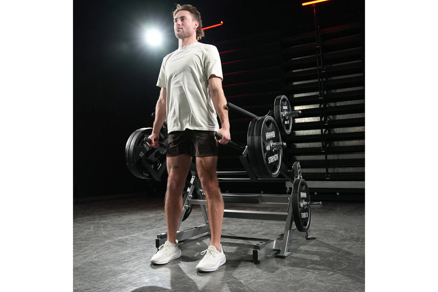 Man wearing light gray t-shirt and black shorts holding dumbbells in gym setting with Hammer Strength equipment