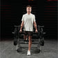 Male model wearing light gray t-shirt and black shorts holding dumbbells in gym setting with Hammer Strength squat machine in
