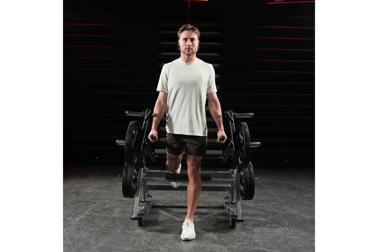 Male model wearing light gray t-shirt and black shorts holding dumbbells in gym setting with Hammer Strength squat machine in