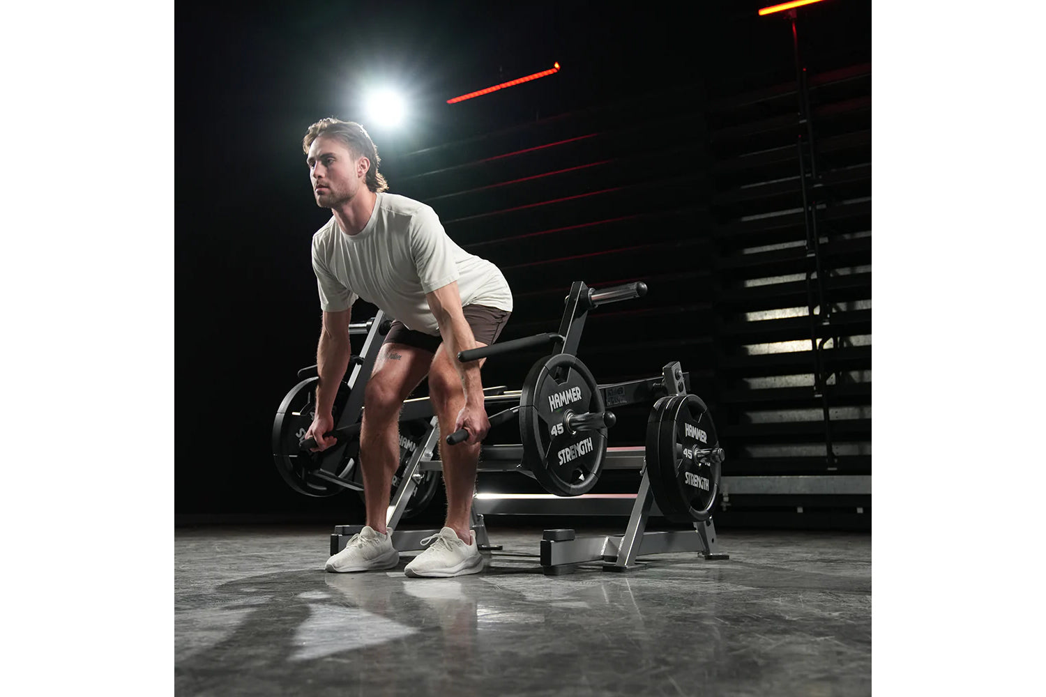 Man in light gray t-shirt and shorts using Hammer Strength multi-squat machine with black weight plates in gym setting with r