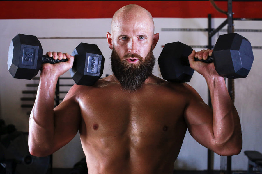 Muscular man holding black hex dumbbells at shoulder height in gym setting