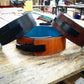 Three leather lever belts in black, cognac brown, and burgundy with contrasting stitching, displayed on wooden work surface