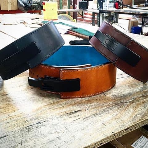 Three leather lever belts in black, cognac brown, and burgundy with contrasting stitching, displayed on wooden work surface