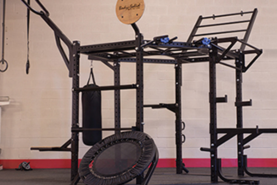 Black multi-function fitness rig with pull-up bar, dip station, and rope attachment, displayed with rebounder trampoline in g