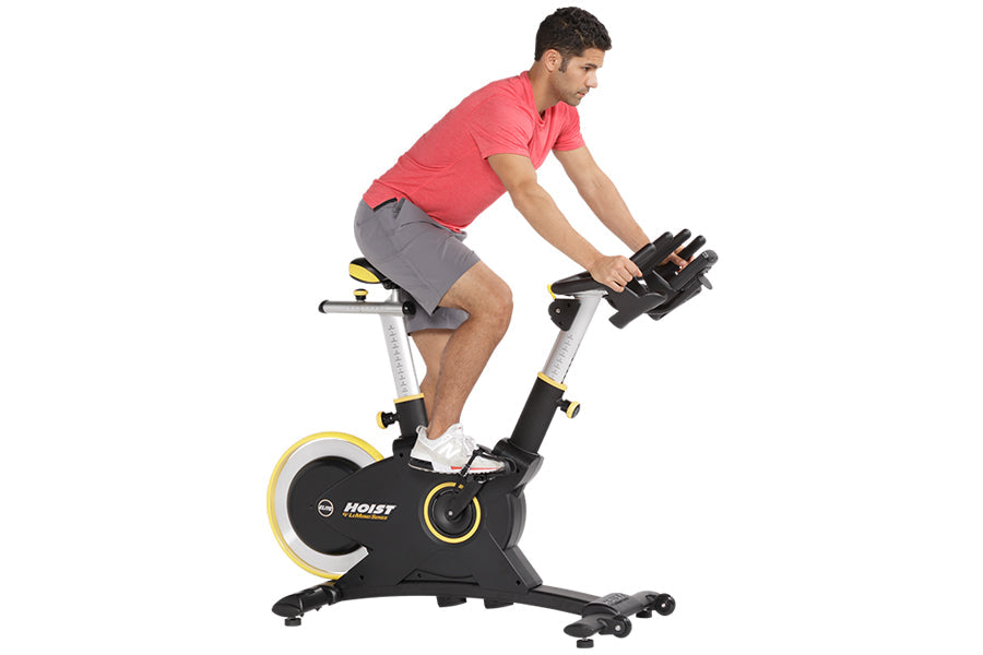 Man in coral t-shirt and gray shorts riding black and yellow Hoist stationary exercise bike with handlebar weights
