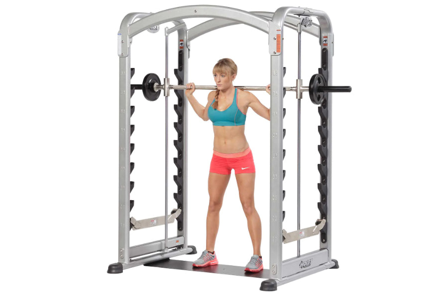 Woman wearing teal sports bra and pink shorts using silver and black Smith machine with barbell for strength training