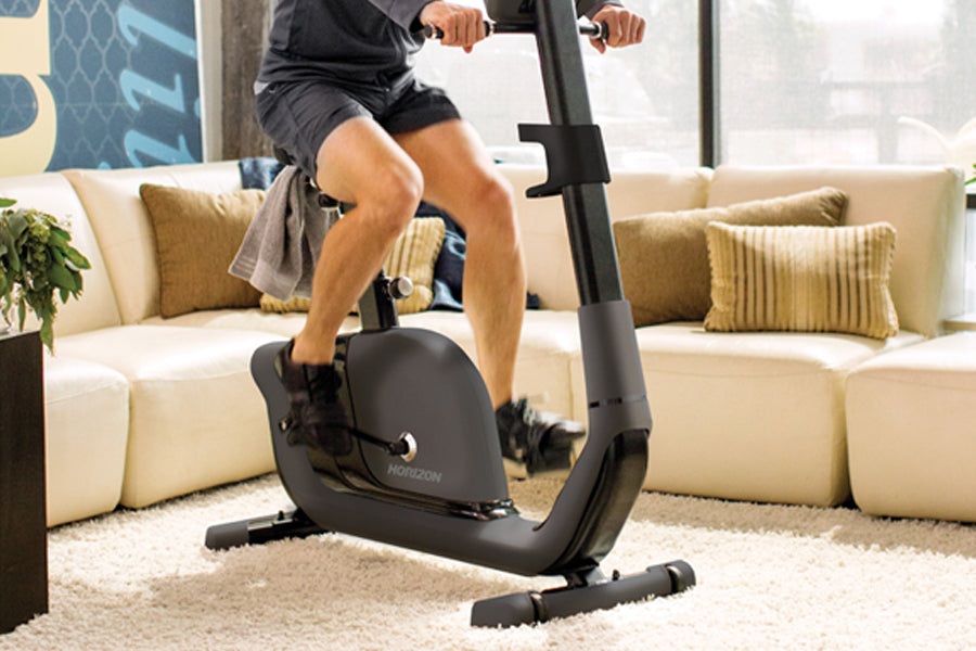 Person using black Horizon upright exercise bike in living room with cream sofa and gold pillows