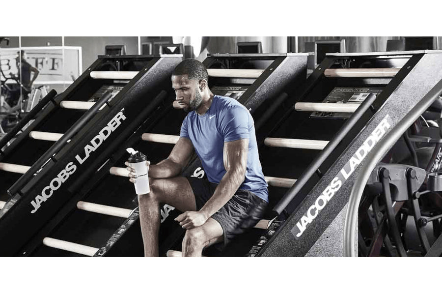 Man in blue t-shirt and black shorts climbing on Jacobs Ladder machine in gym setting, holding white water bottle