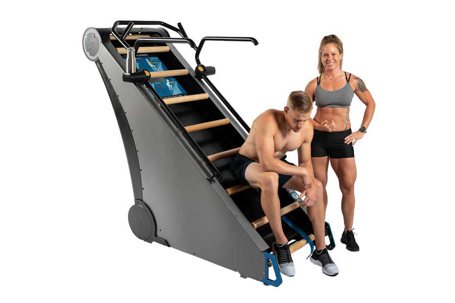 Gray and black stair climbing machine with wooden steps and blue accents, shown with two people demonstrating use on white ba