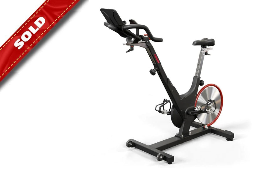 Black and red indoor exercise bike with upright frame, adjustable seat and handlebars, red flywheel, on white background