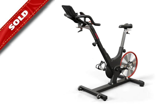Black and red indoor exercise bike with upright frame, adjustable seat and handlebars, red flywheel, on white background