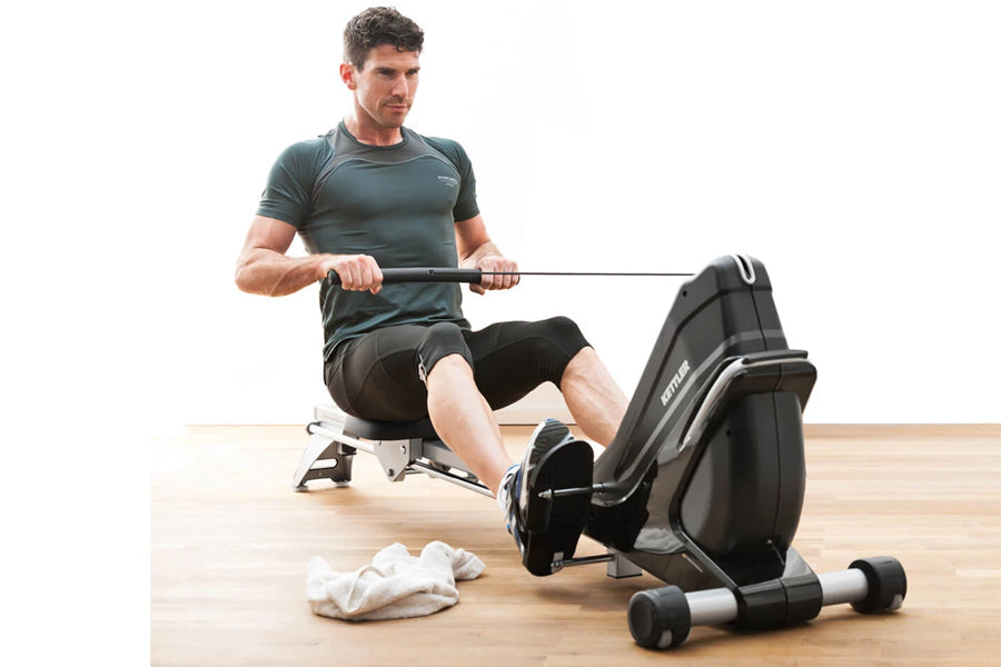 Man using black Kettler Coach E rowing machine, wearing gray t-shirt and black shorts on wooden floor