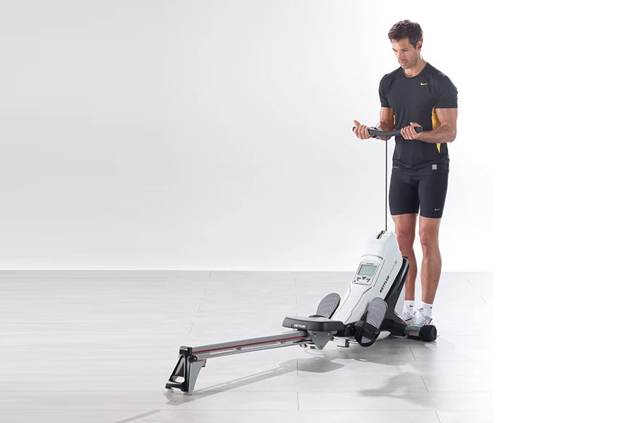 Man using black Kettler Coach E rowing machine on white background, wearing dark t-shirt and shorts