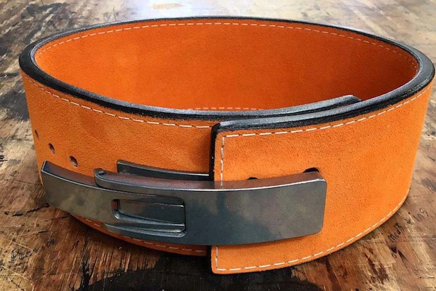 Orange suede weightlifting belt with black lever buckle and white stitching, displayed on wooden surface