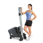 Woman in blue tank top and black shorts demonstrating upright stationary exercise bike with vertical silver column and black 
