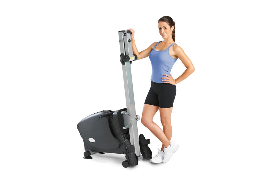 Woman in blue tank top and black shorts demonstrating upright stationary exercise bike with vertical silver column and black 