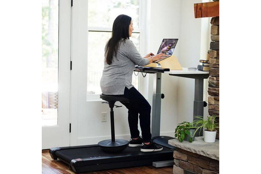 Woman in gray sweater and black pants using LifeSpan TR1000-DT7 treadmill desk with laptop in bright office space