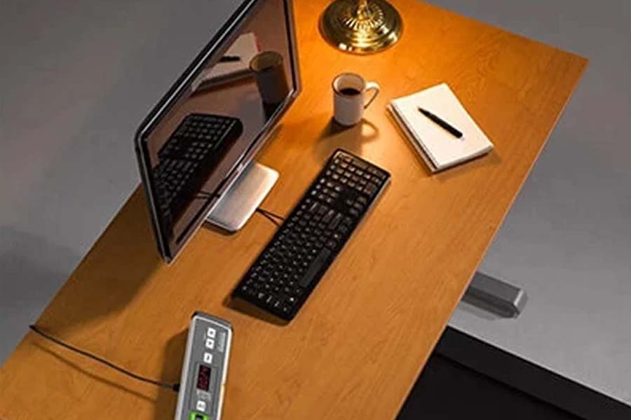 Overhead view of wooden desk with black keyboard, monitor, ceramic mug, notepad, brass lamp, and power strip showing under-de
