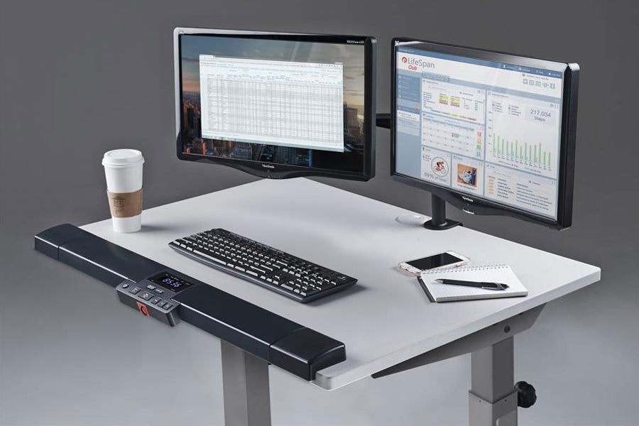 White desktop treadmill with black motorized base, dual monitors displaying spreadsheets, wireless keyboard, coffee cup, and 