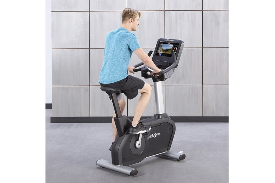 Man in blue t-shirt and black shorts riding black and silver upright stationary bike with digital display console, gray tile 