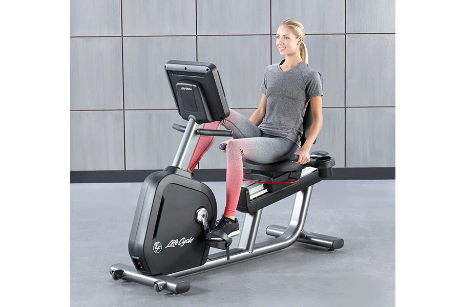 Woman in gray t-shirt and pink leggings seated on Life Fitness recumbent exercise bike with digital display, silver frame, an