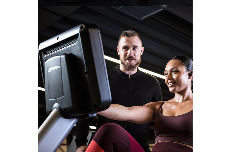 Two people using black recumbent exercise bike in modern gym setting with overhead lighting