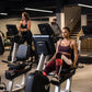 Woman in burgundy sports bra and leggings on recumbent stationary bike in gym setting with multiple cardio machines