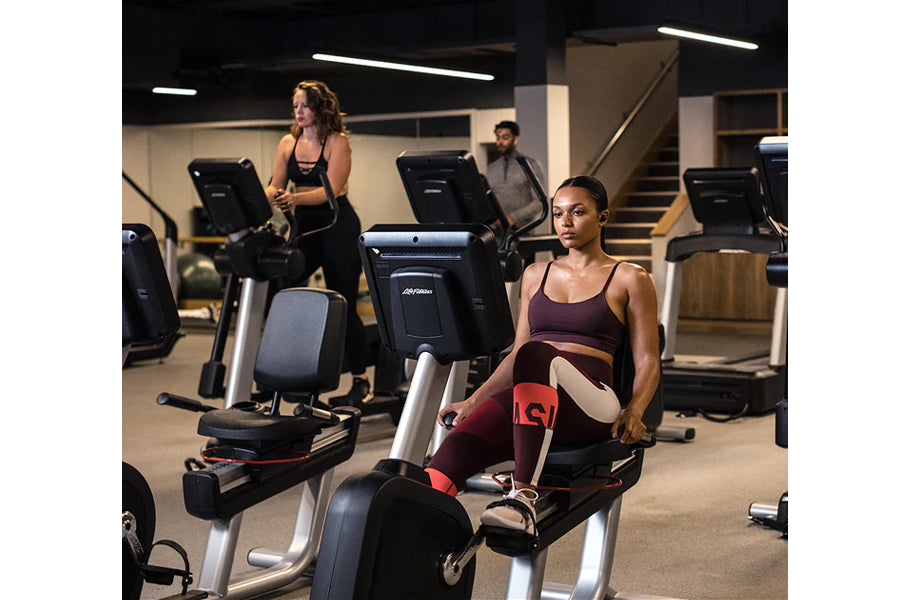 Woman in burgundy sports bra and leggings on recumbent stationary bike in gym setting with multiple cardio machines