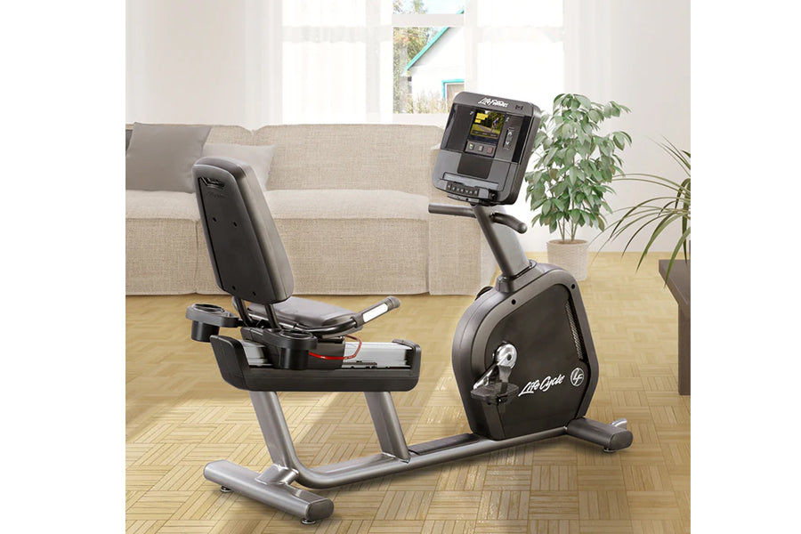 Black and silver recumbent exercise bike with padded seat and monitor display, positioned in living room setting with beige s