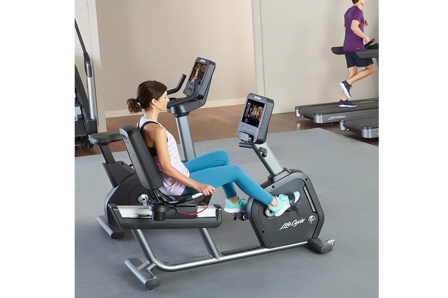 Woman in white tank top and blue leggings using black Life Fitness recumbent exercise bike with digital display in gym settin