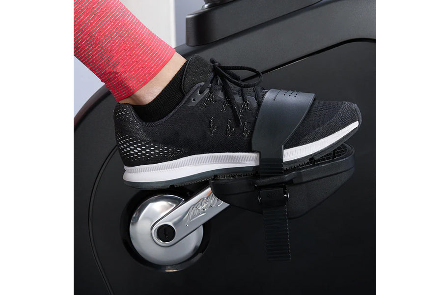 Black mesh athletic shoe with white sole stripe worn by model on exercise bike pedal, red ribbed sweater visible above