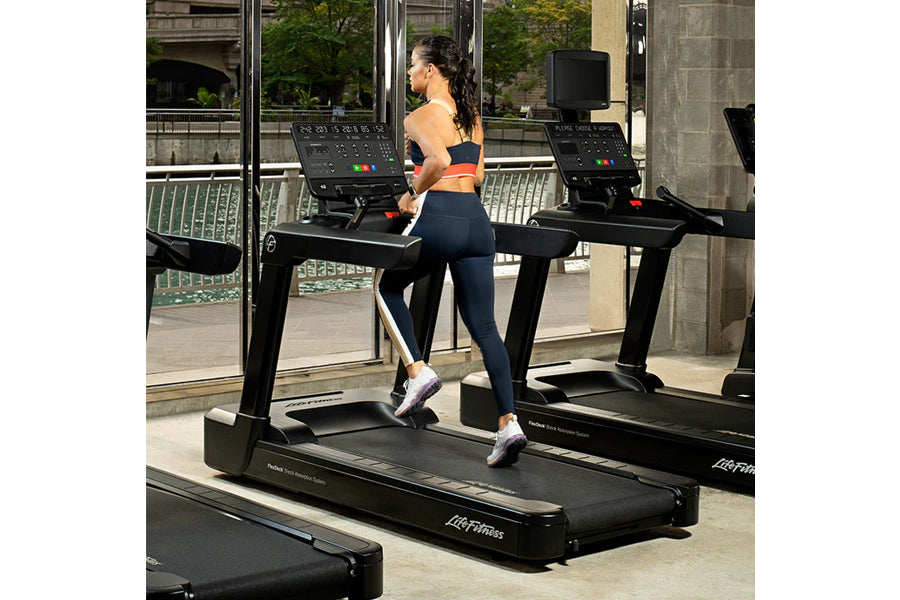 Woman running on black Life Fitness treadmill in gym, wearing navy leggings and orange sports bra
