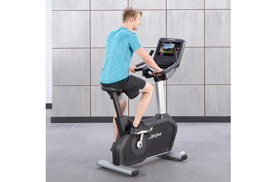 Man wearing turquoise t-shirt and black shorts exercising on black and silver upright stationary bike with digital display co
