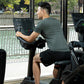 Man in gray shirt and black shorts using upright stationary bike with digital console display in gym overlooking park