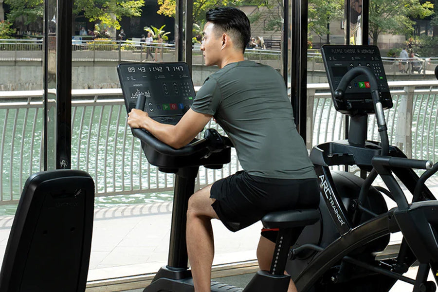 Man in gray shirt and black shorts using Life Fitness upright exercise bike with digital display console in gym setting near 