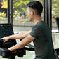 Man in dark gray t-shirt using upright stationary exercise bike with digital console display in bright gym setting with windo