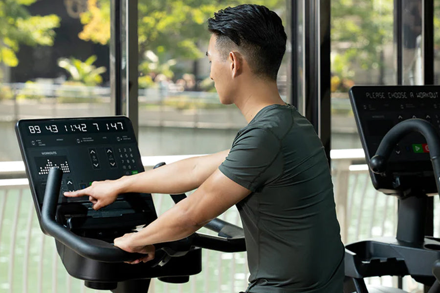 Man in dark gray t-shirt using upright stationary exercise bike with digital console display in bright gym setting with windo