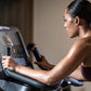 Woman using black Life Fitness upright stationary bike with digital console display, wearing burgundy sports bra