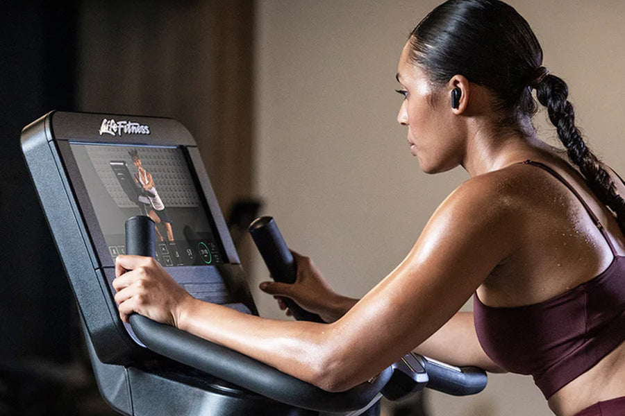 Woman in burgundy sports bra using upright stationary bike with digital console display, side view