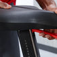 Gray padded seat with black base held by hands above stationary bike upright, worn by model in red shirt