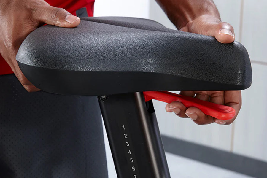 Hands adjusting gray padded seat on upright exercise bike with numbered resistance levels, worn by model in red shirt