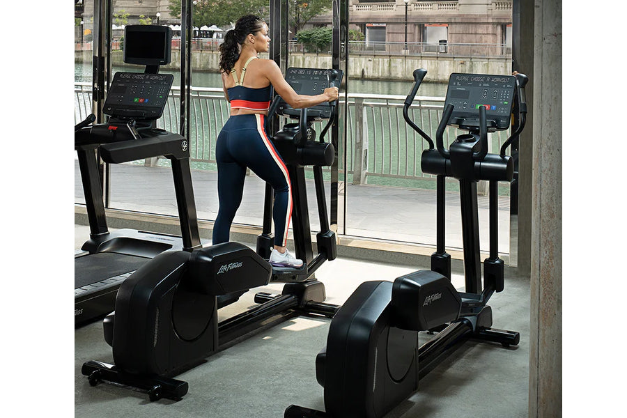 Woman exercising on black Life Fitness elliptical trainer wearing red and navy athletic wear in modern gym studio with window