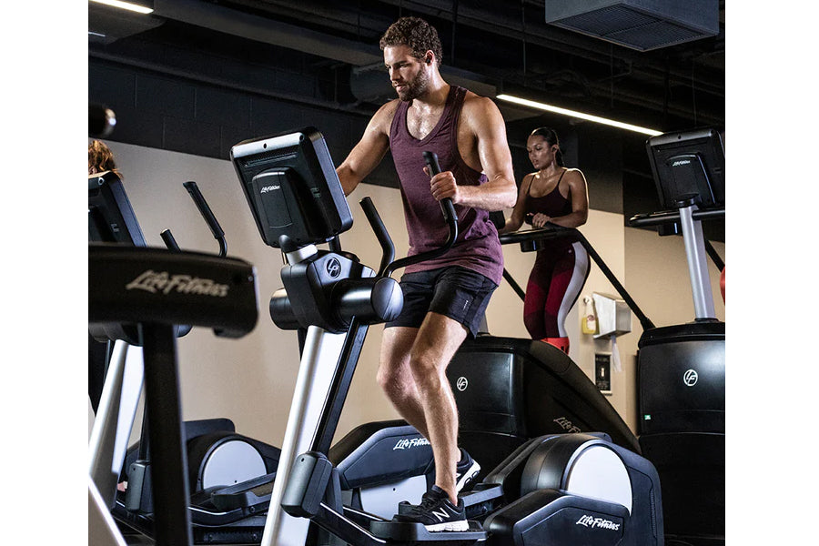 Man wearing burgundy tank top and black shorts exercising on Life Fitness elliptical machine in gym setting with other equipm