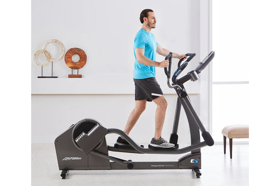 Man in blue t-shirt using black and white Life Fitness E5 elliptical machine in bright fitness studio setting