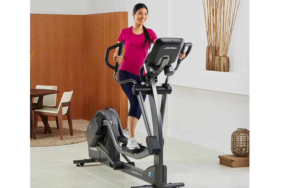 Woman in magenta t-shirt using black and gray Life Fitness elliptical machine with digital display in home gym setting