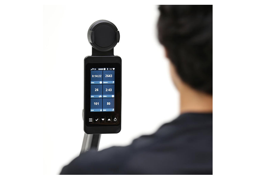Black digital fitness monitor with touchscreen display showing workout metrics, mounted on rowing machine pole, held by perso